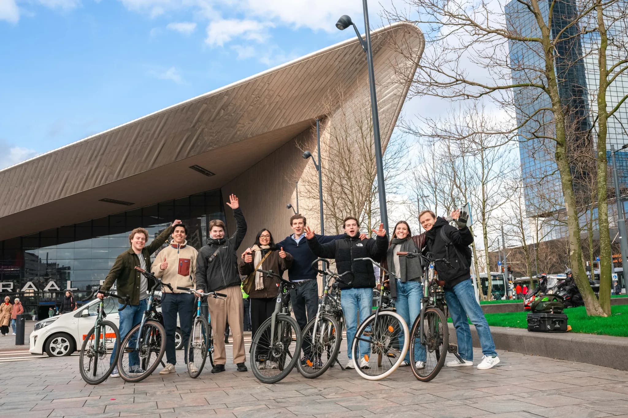 BIKEBAZE-teamleden zwaaien naast hun fietsen voor het golvende dak van Rotterdam Centraal.