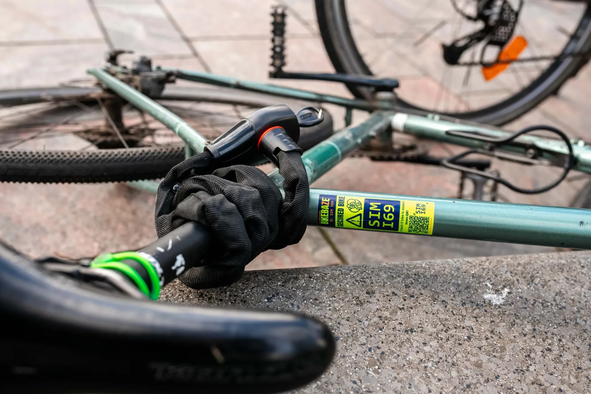 Toppled bike with chain lock and yellow BIKEBAZE SafeTag on green frame beside curb.