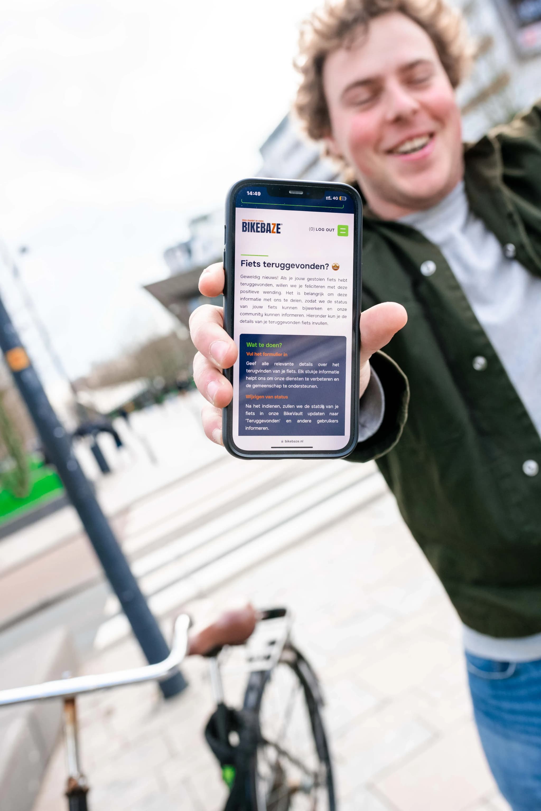 Lachende man toont smartphone met BIKEBAZE pagina 'Fiets teruggevonden' op straat naast zijn fiets.