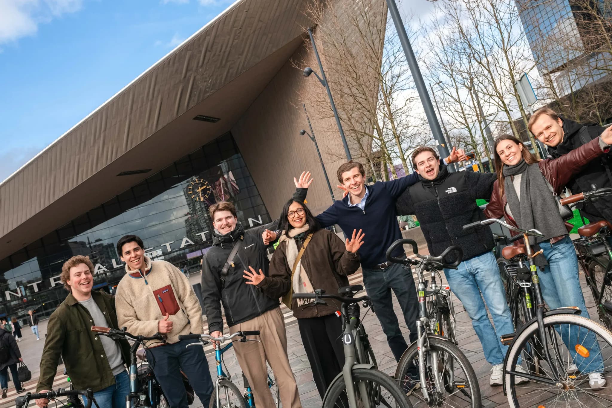 Group of smiling young people with bikes in front of Rotterdam Centraal Station on a sunny day.