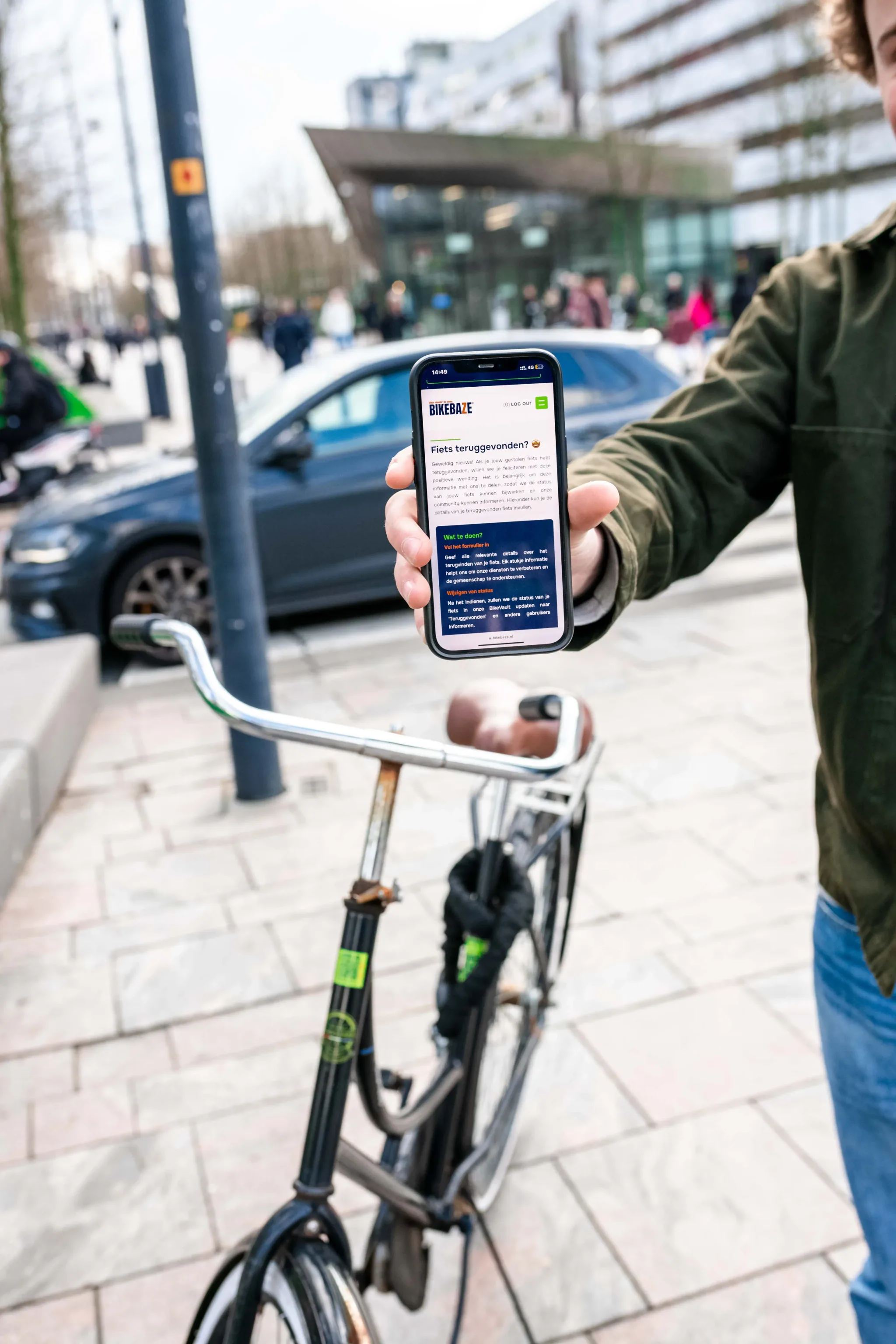 Hand toont smartphone met BIKEBAZE-melding boven stuur van fiets op stadsplein.