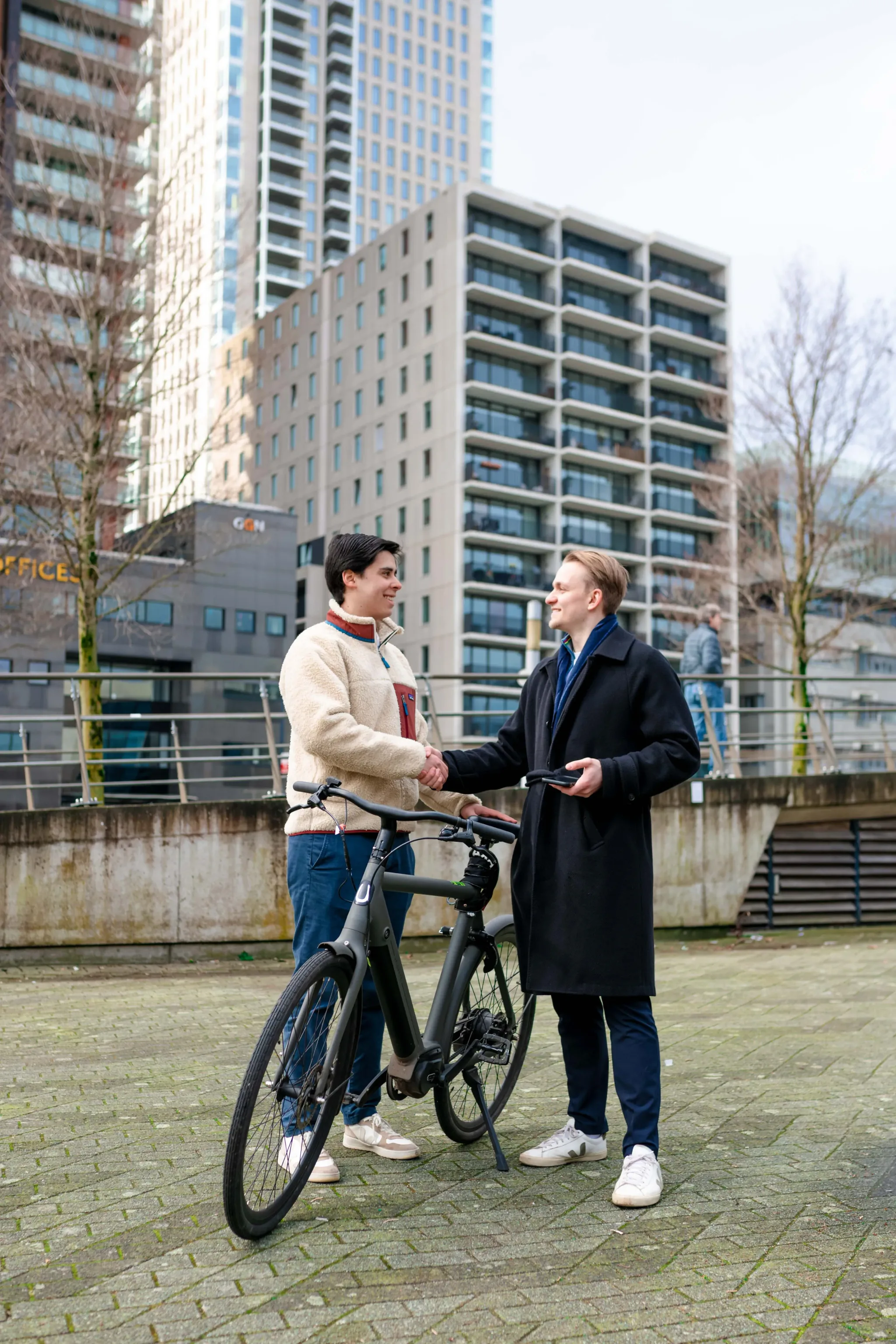 Twee jonge mannen schudden elkaar de hand naast een fiets in een Rotterdamse stadsomgeving.