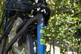 Blauwe BIKEBAZE SafeTag-sticker met QR-code op zwarte voorvork tussen groen blad.