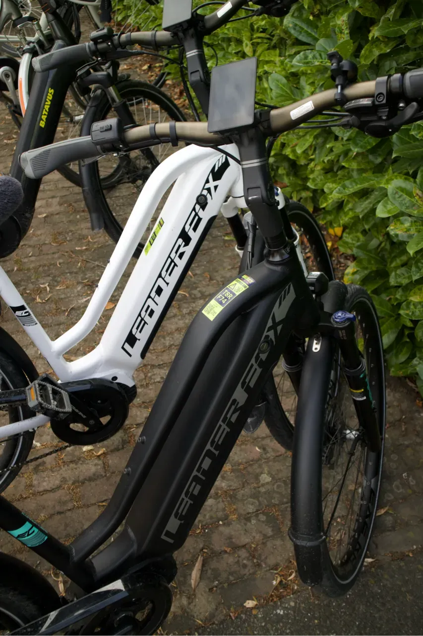 White and black Leader Fox e-bikes with BIKEBAZE SafeTag stickers parked beside green hedge.