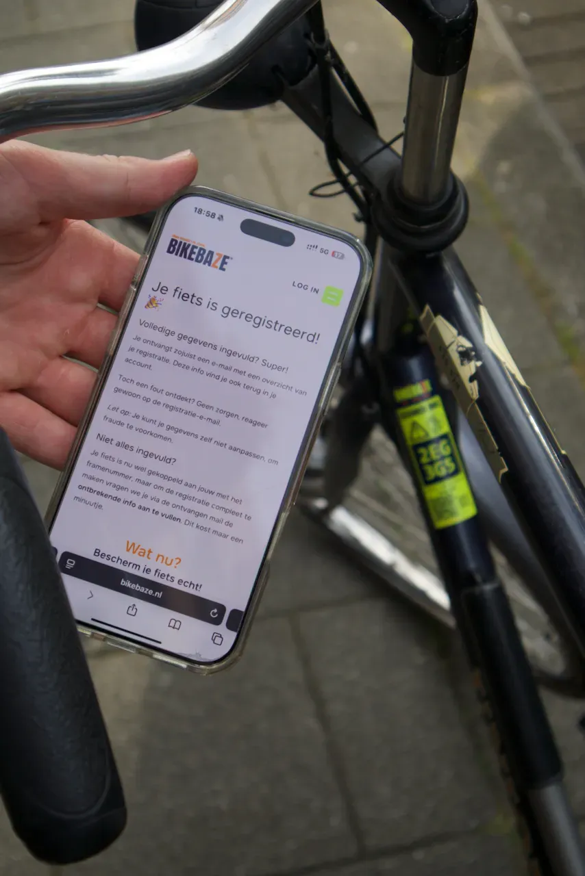 Hand toont smartphone met BIKEBAZE registratiebevestiging naast fiets met gele SafeTag op het frame.