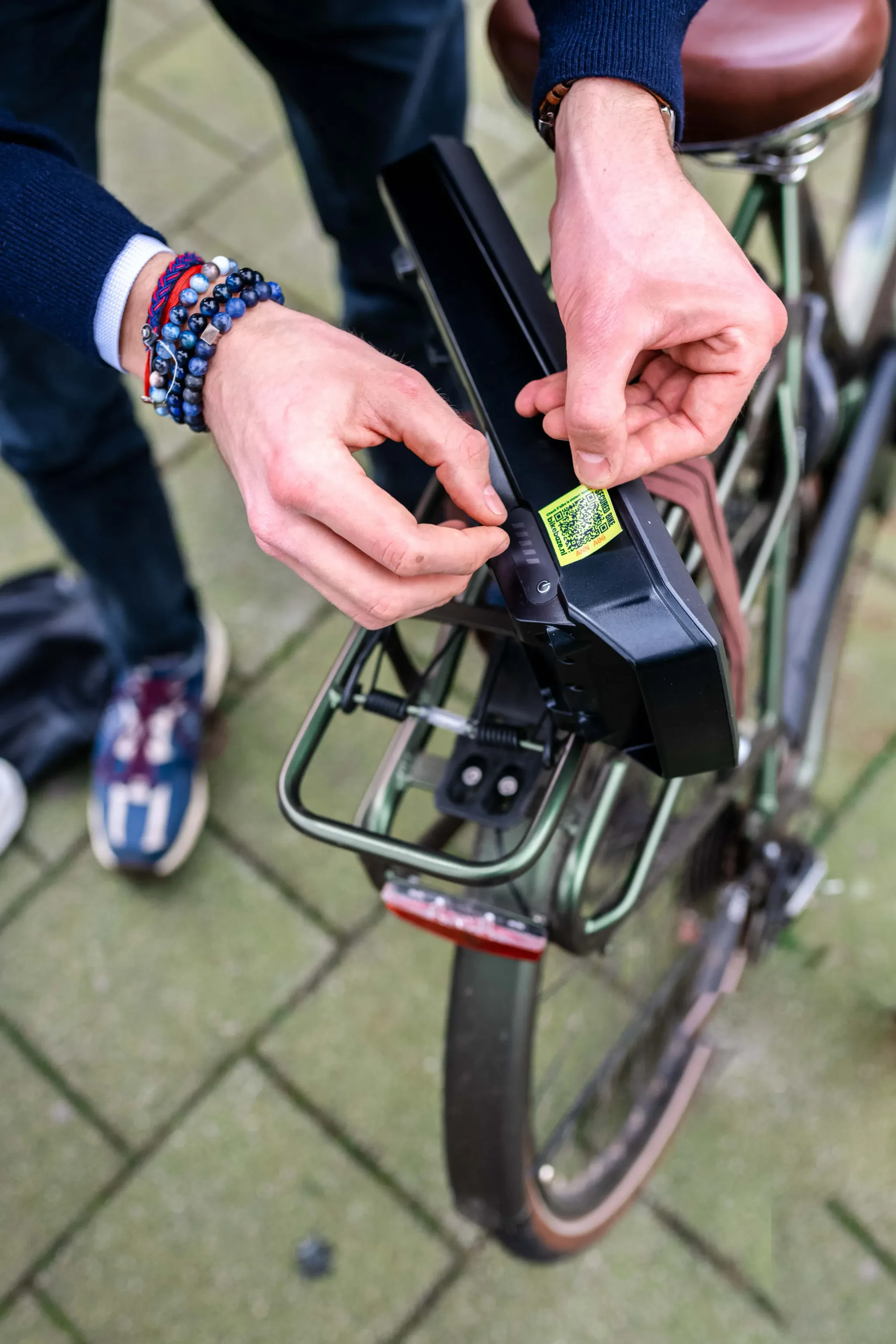 Handen bevestigen een gele BIKEBAZE SafeTag op de bagagedrager-accu van een groene fiets.