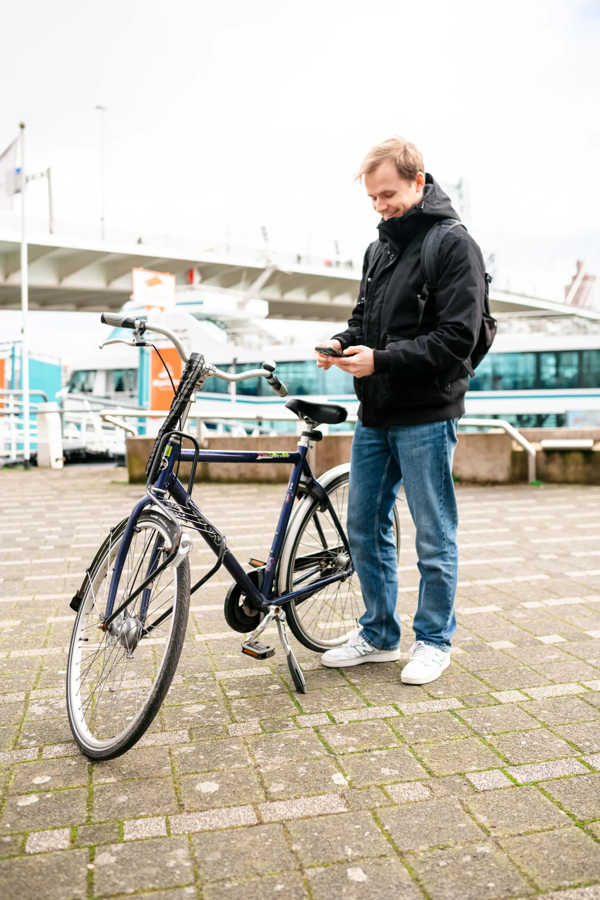 Man in zwarte jas bekijkt zijn telefoon naast een donkerblauwe stadsfiets op de Rotterdamse kade.