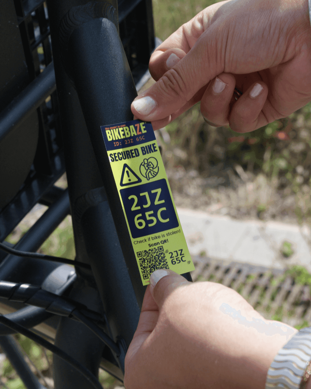 Two hands applying a yellow BIKEBAZE SafeTag with QR code and code 2JZ 65C onto a black bike frame.