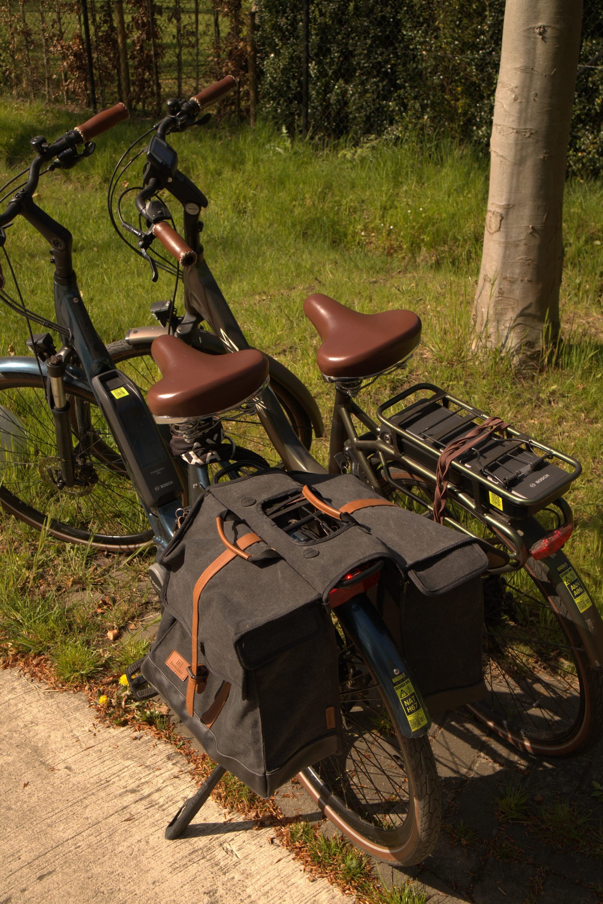 Twee e-bikes met bruine zadels en zwarte fietstassen geparkeerd op een tuinpad in de zon.