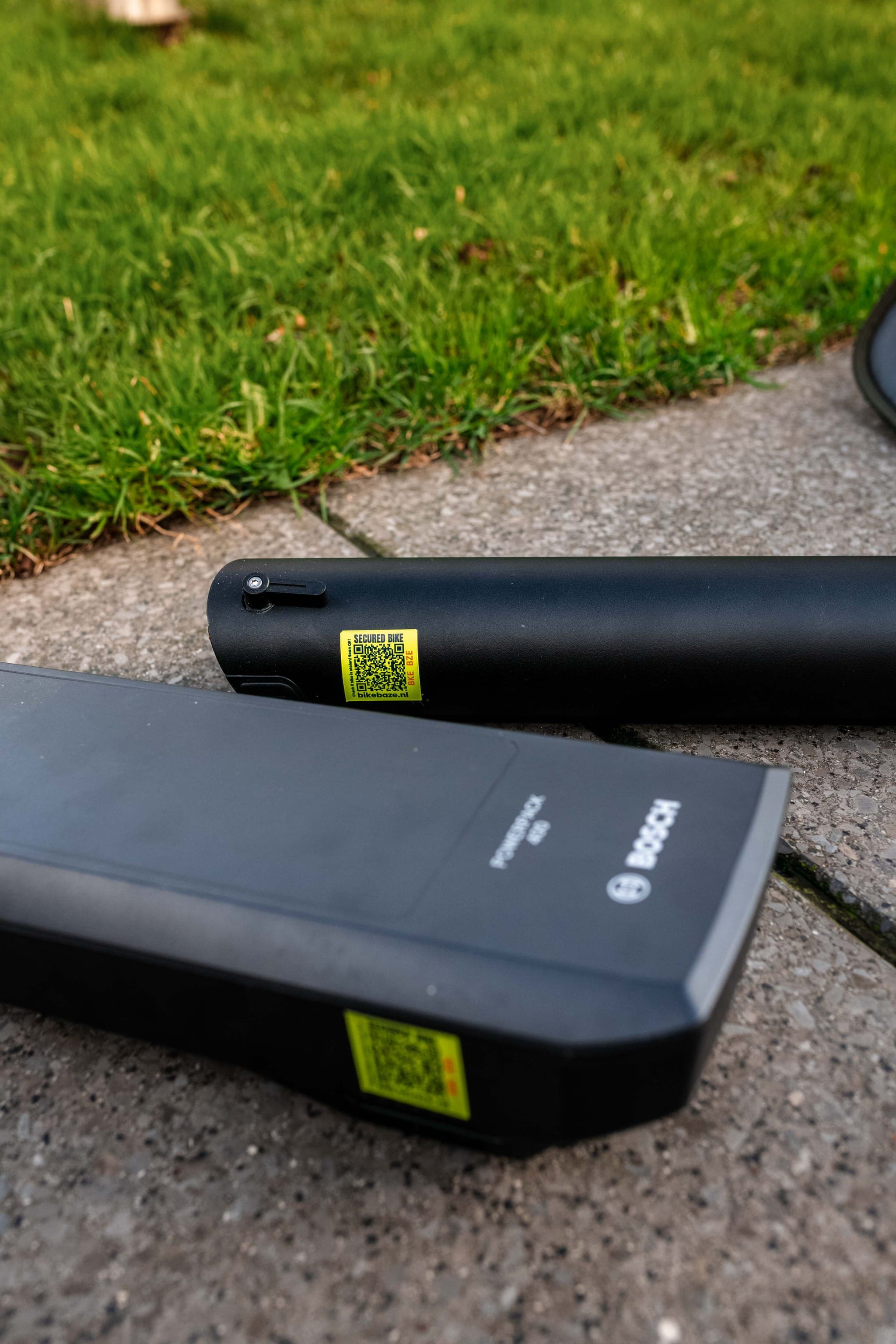 Twee zwarte e-bike accu's, waaronder een Bosch model, liggen op tegels naast gras met gele BIKEBAZE SafeTag QR-stickers.