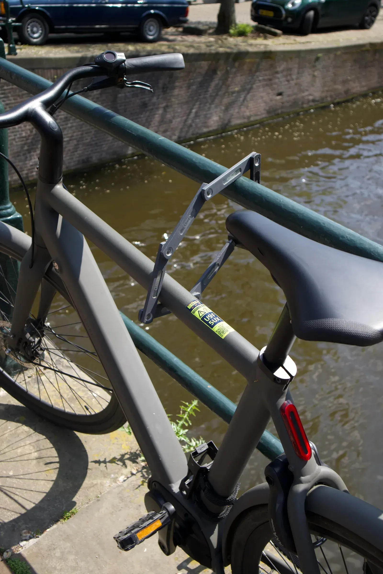 Grijze e-bike met vouwslot aan een groene reling langs een Amsterdamse gracht, met een BIKEBAZE SafeTag op het frame.